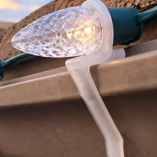 Detail: a clear plastic clip hooked over the edge of a gutter, holding a warm-white C9 bulb
