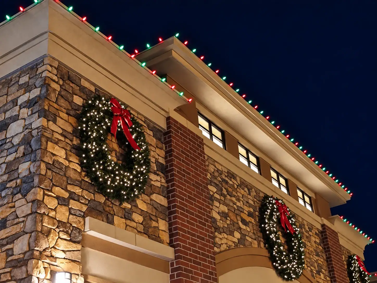 Stone and brick commercial building with red, green and white roofline lights and lit wreaths at night