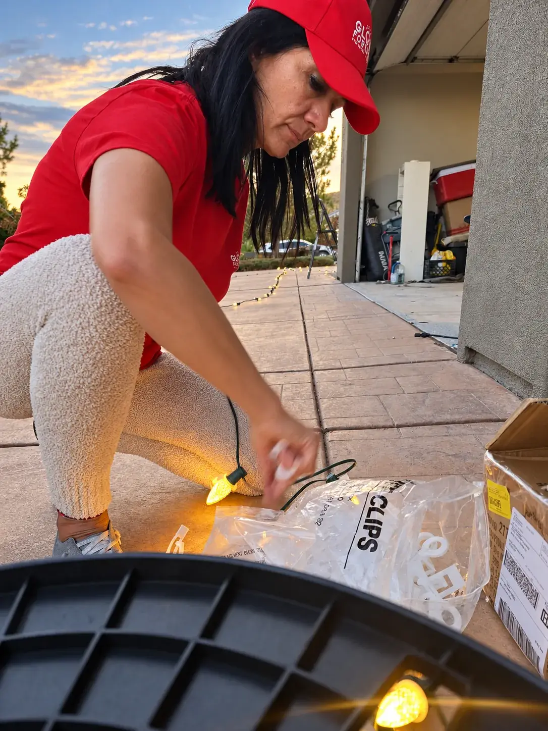 Glow Forward crew member in a red branded cap and shirt sorting light clips and bulbs by a garage at dusk