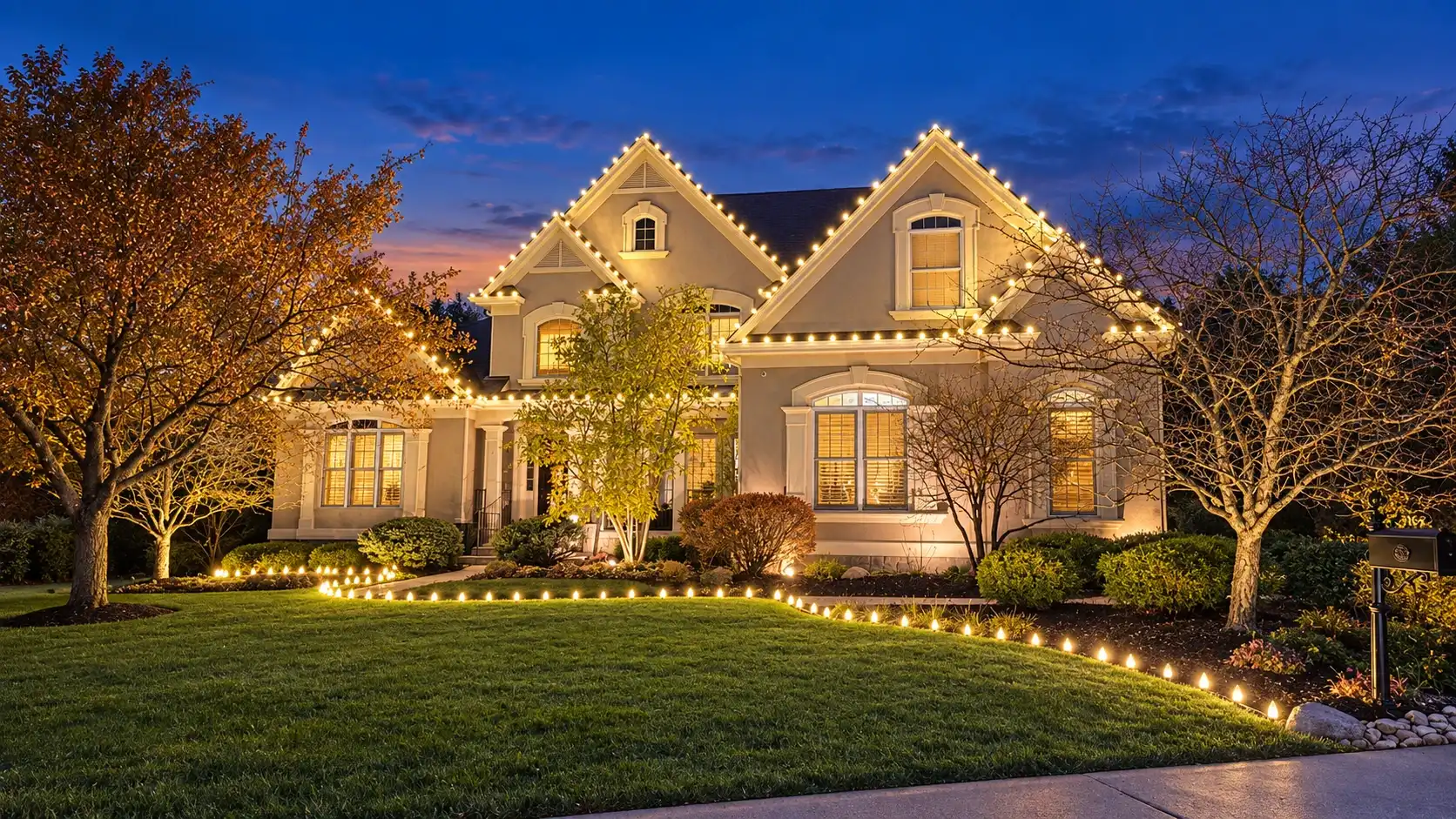 Two-story home with warm-white Christmas roofline lights and a lit pathway border at dusk