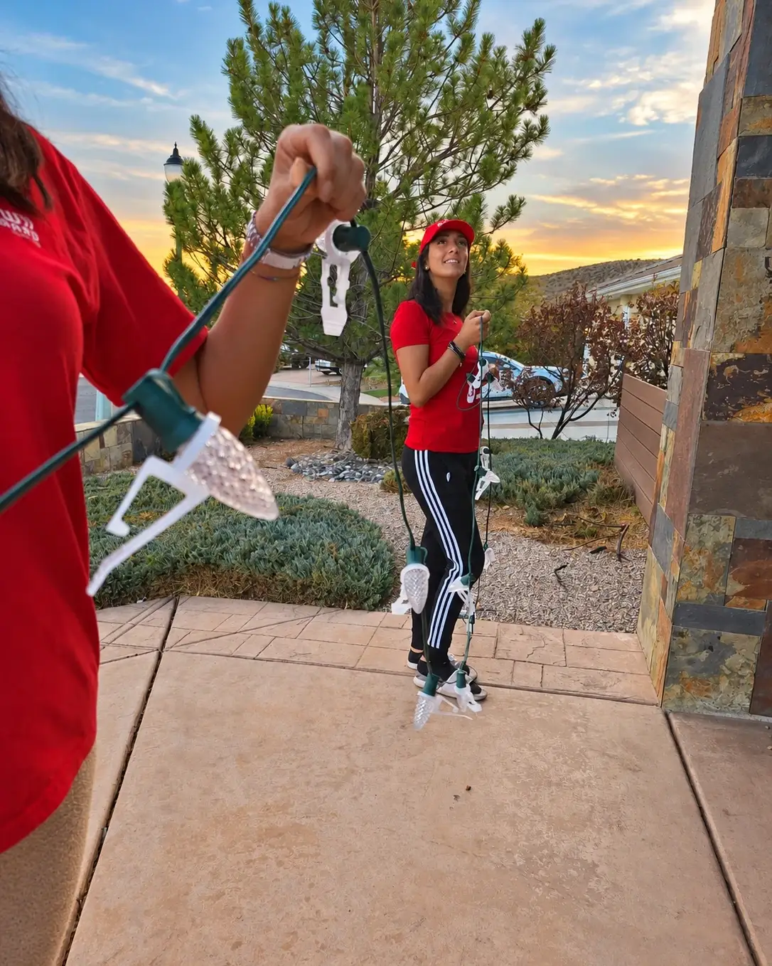 Glow Forward crew member in a red branded cap holding a strand of C9 bulbs on clips beside a home at sunset, a teammate holding the strand behind
